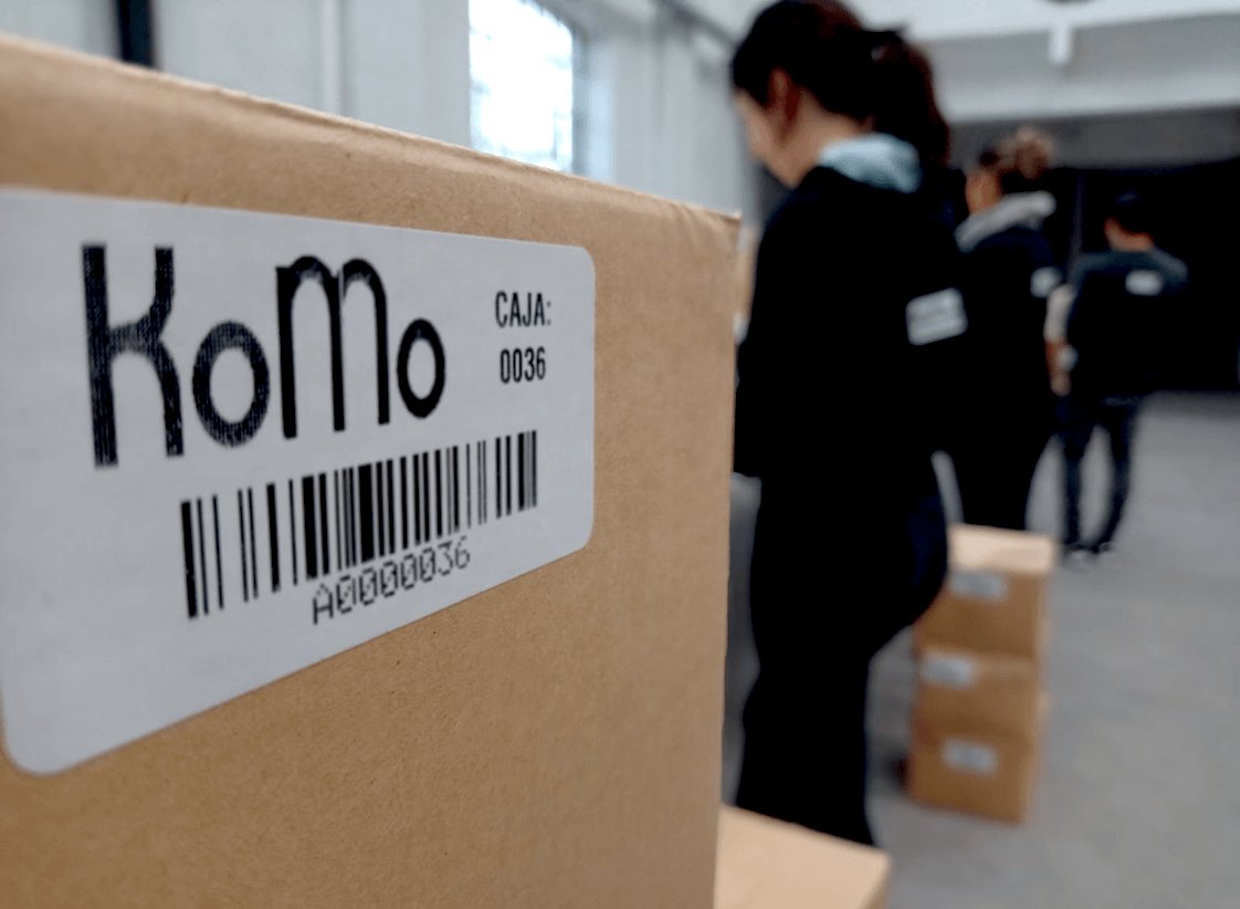 Caja de archivo KoMo etiquetada con código de barras y operarios de planta trabajando al fondo